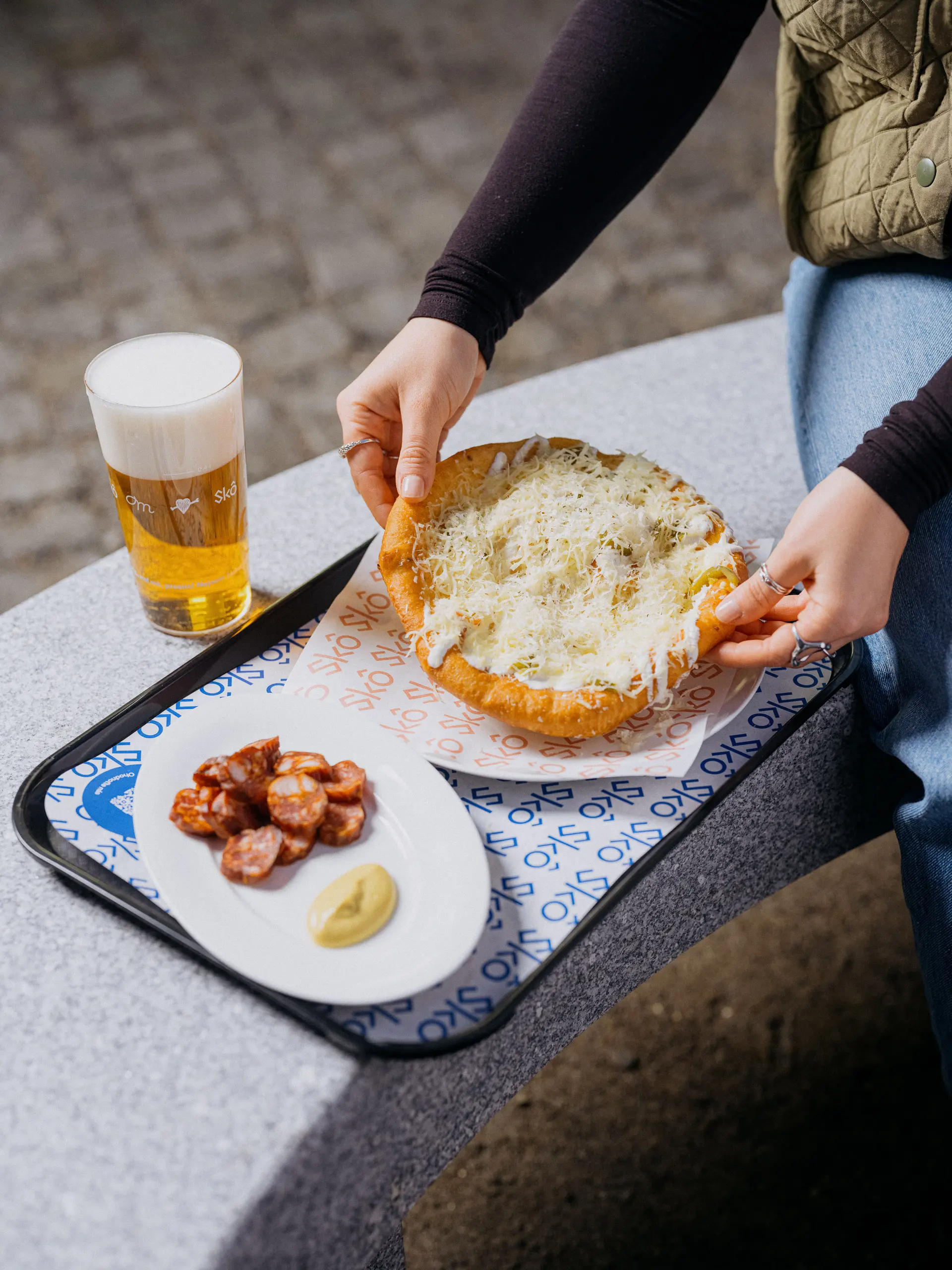Langoš se sýrem a klobása s hořčicí na podnosu ve Skô, pivo vedle.